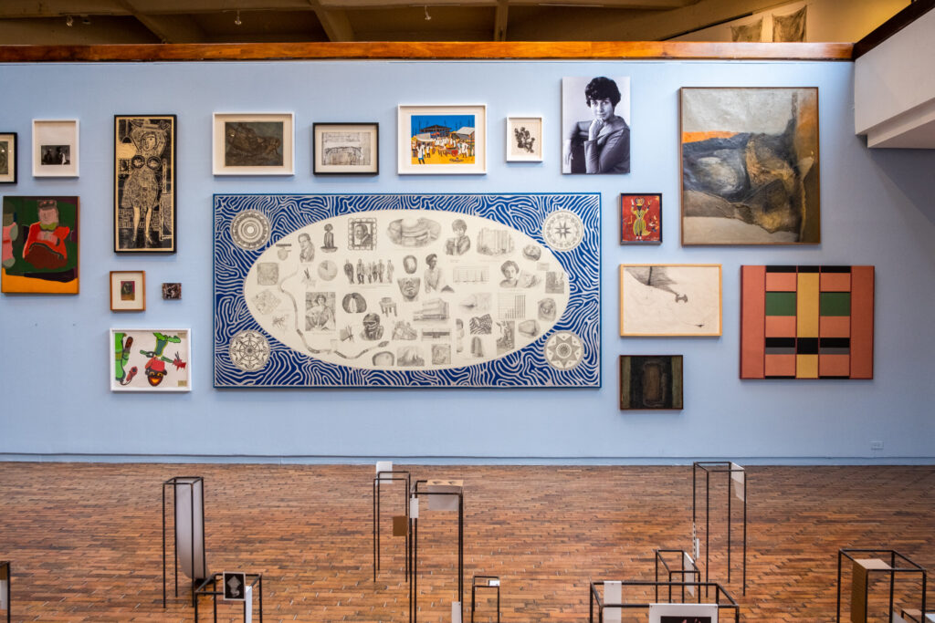 Vistas de sala de la exposición Viceversa: posiciones y perspectivas de una colección (2023) en el Museo de Arte Moderno de Bogotá. En la pared azul, arriba a la derecha, la obra Disonancia sentimental (1961) de Maruja Rolando. Tomado de: https://www.mambogota.com/actividad/adriana-bustos-proyecto-de-sitio-especifico/ y https://www.mambogota.com/wp-content/uploads/2023/12/mambo-Viceversa-I.V-Piso-2-GDzg-62-min-scaled.jpg