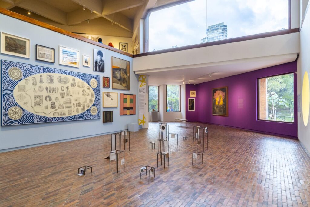 Vistas de sala de la exposición Viceversa: posiciones y perspectivas de una colección (2023) en el Museo de Arte Moderno de Bogotá. En la pared azul, arriba a la derecha, la obra Disonancia sentimental (1961) de Maruja Rolando. Tomado de: https://www.mambogota.com/actividad/adriana-bustos-proyecto-de-sitio-especifico/ y https://www.mambogota.com/wp-content/uploads/2023/12/mambo-Viceversa-I.V-Piso-2-GDzg-62-min-scaled.jpg